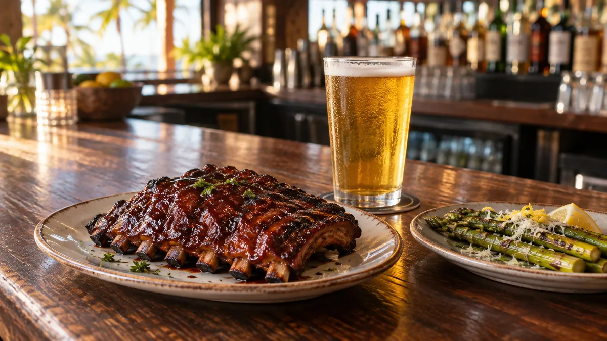 A glazed rack of baby back ribs and a glass of pale draft lager on a bar counter in late-afternoon light
