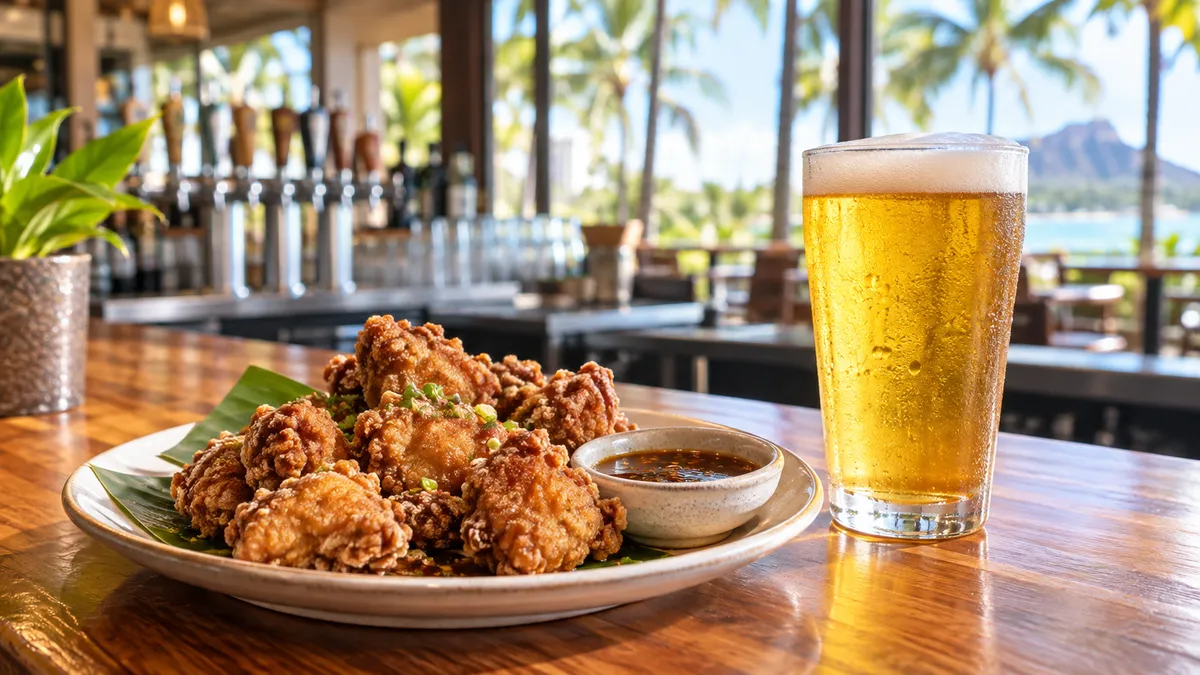 A plate of crispy fried chicken and a pint of pale beer on a wooden bar table in soft mid-afternoon light, with palm shadows on the wall
