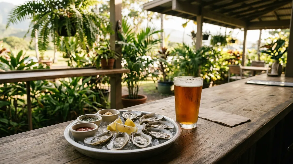 A tray of oysters on the half shell on crushed ice with lemon wedges and small sauce cups, next to a pint of IPA on a bar counter surrounded by green plants in late-afternoon light