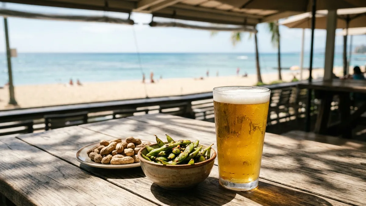 An open-air beachfront restaurant table in late-afternoon light with a pint of golden ale, a bowl of edamame and a small dish of boiled peanuts, the ocean and a pale sandy beach beyond