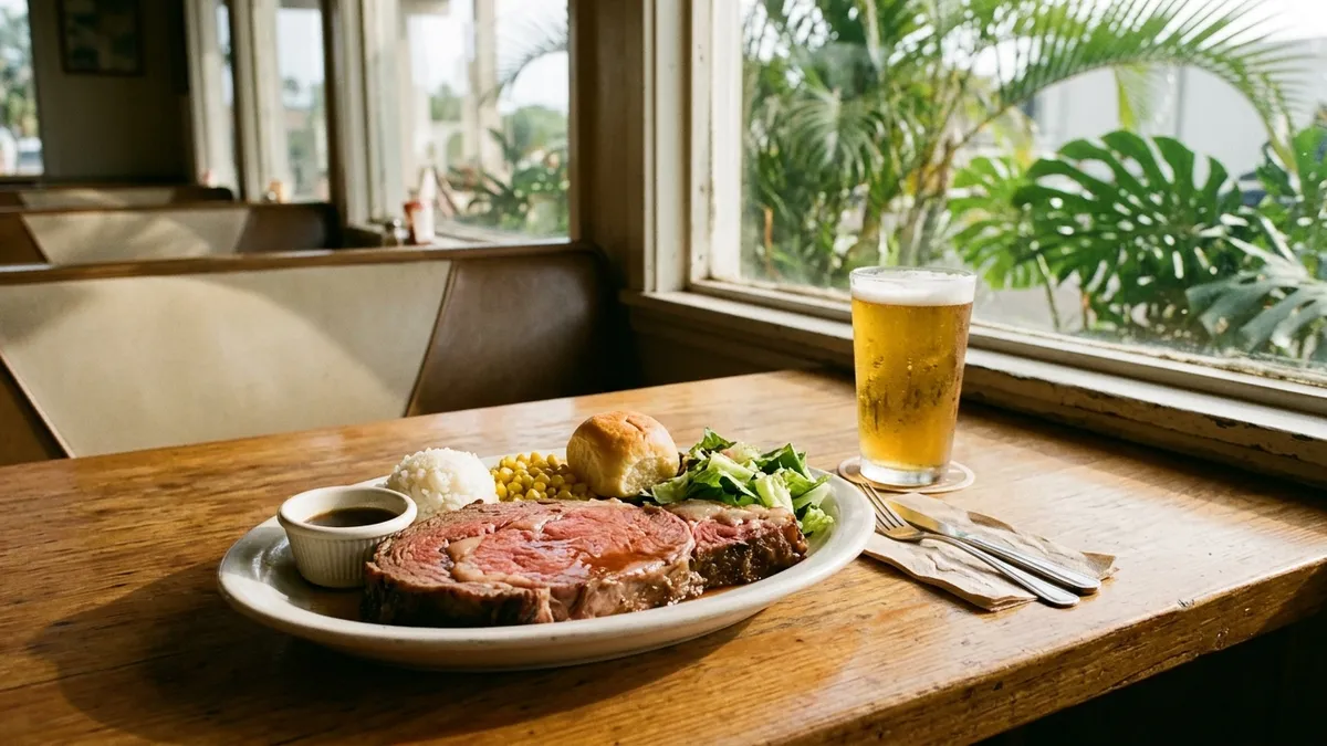 A plate of sliced prime rib with rice, corn and a dinner roll next to a glass of beer on a restaurant table in late-afternoon light