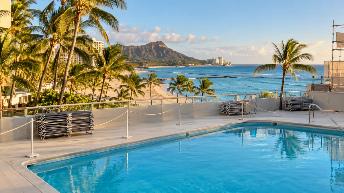 A beachfront hotel on Waikiki Beach in late-afternoon light, with an empty pool deck closed off by a simple rope line in the foreground and the ocean beyond