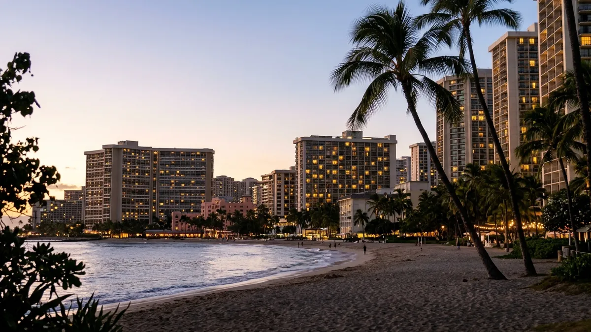 Waikiki hotel towers at early dusk seen from the beach side, some windows lit and some dark, palm trees and a calm ocean in the foreground